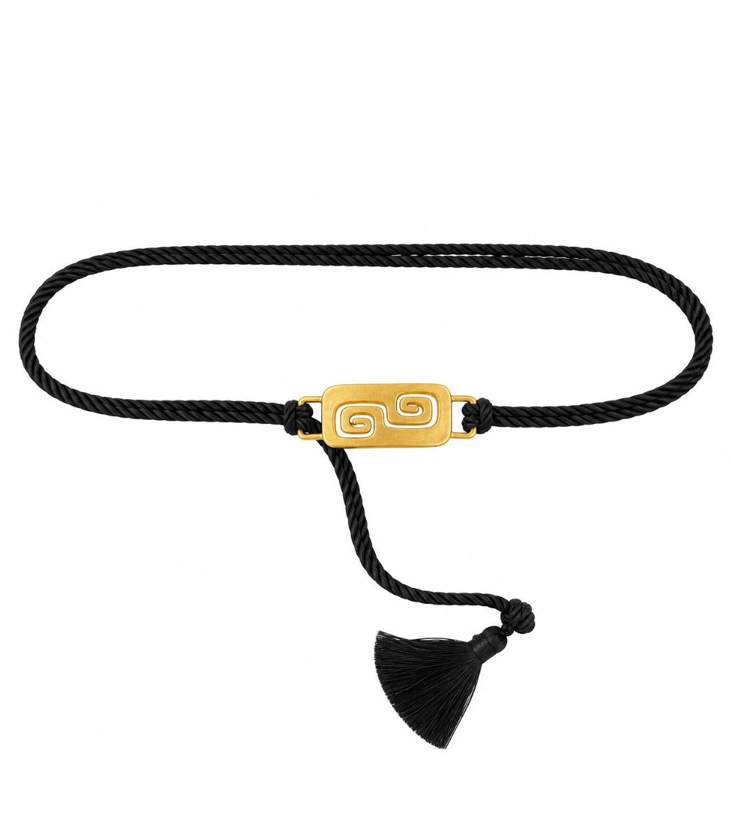 A black braided cord bracelet with a polished gold rectangular plate featuring a Greek key swirl design, finished with a black tassel.