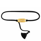 A black braided cord bracelet with a polished gold rectangular plate featuring a Greek key swirl design, finished with a black tassel.