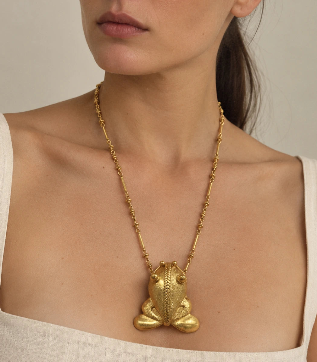 A close-up of a model wearing a gold necklace with a textured frog pendant, styled with a white top against a neutral beige background.
