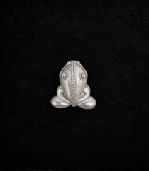 A silver frog-shaped brooch with detailed textured engravings, centered on a textured black background.