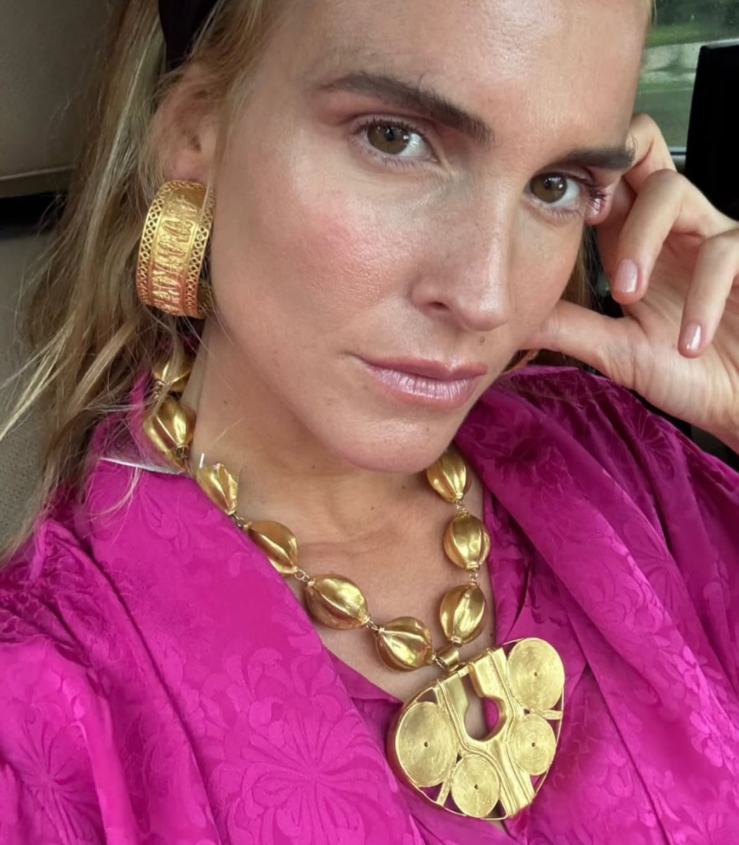 A close-up lifestyle shot of a woman wearing a chunky gold statement necklace and matching textured gold hoop earring, paired with a vibrant magenta top.