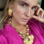 A close-up lifestyle shot of a woman wearing a chunky gold statement necklace and matching textured gold hoop earring, paired with a vibrant magenta top.