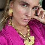 A close-up lifestyle shot of a woman wearing a chunky gold statement necklace and matching textured gold hoop earring, paired with a vibrant magenta top.