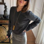 A woman wearing a dark gray V-neck knit sweater and a gray skirt, accessorized with a long gold pendant necklace and black sunglasses, posing indoors next to an ornate cabinet.