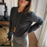 A woman wearing a dark gray V-neck knit sweater and a gray skirt, accessorized with a long gold pendant necklace and black sunglasses, posing indoors next to an ornate cabinet.