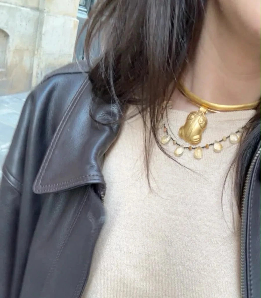 A close-up of a person wearing layered gold necklaces, including a thick choker and a delicate chain with a frog pendant, styled with a black leather jacket over a beige top.