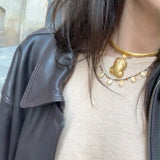 A close-up of a person wearing layered gold necklaces, including a thick choker and a delicate chain with a frog pendant, styled with a black leather jacket over a beige top.
