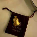 A gold pendant necklace displayed on a dark brown velvet drawstring pouch with the brand name CANO and 'Since 1970' printed on it, set against a light beige background.