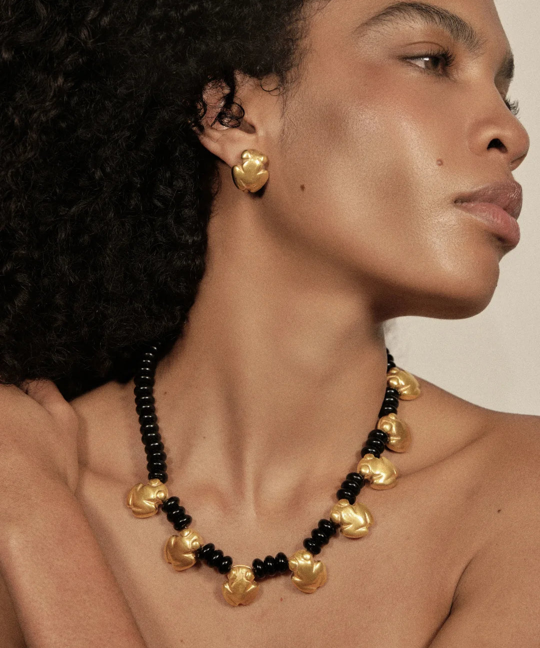 A close-up of a model wearing a statement necklace and matching stud earring. The jewelry features textured, organic-shaped gold beads alternating with small glossy black beads, set against a warm neutral background.