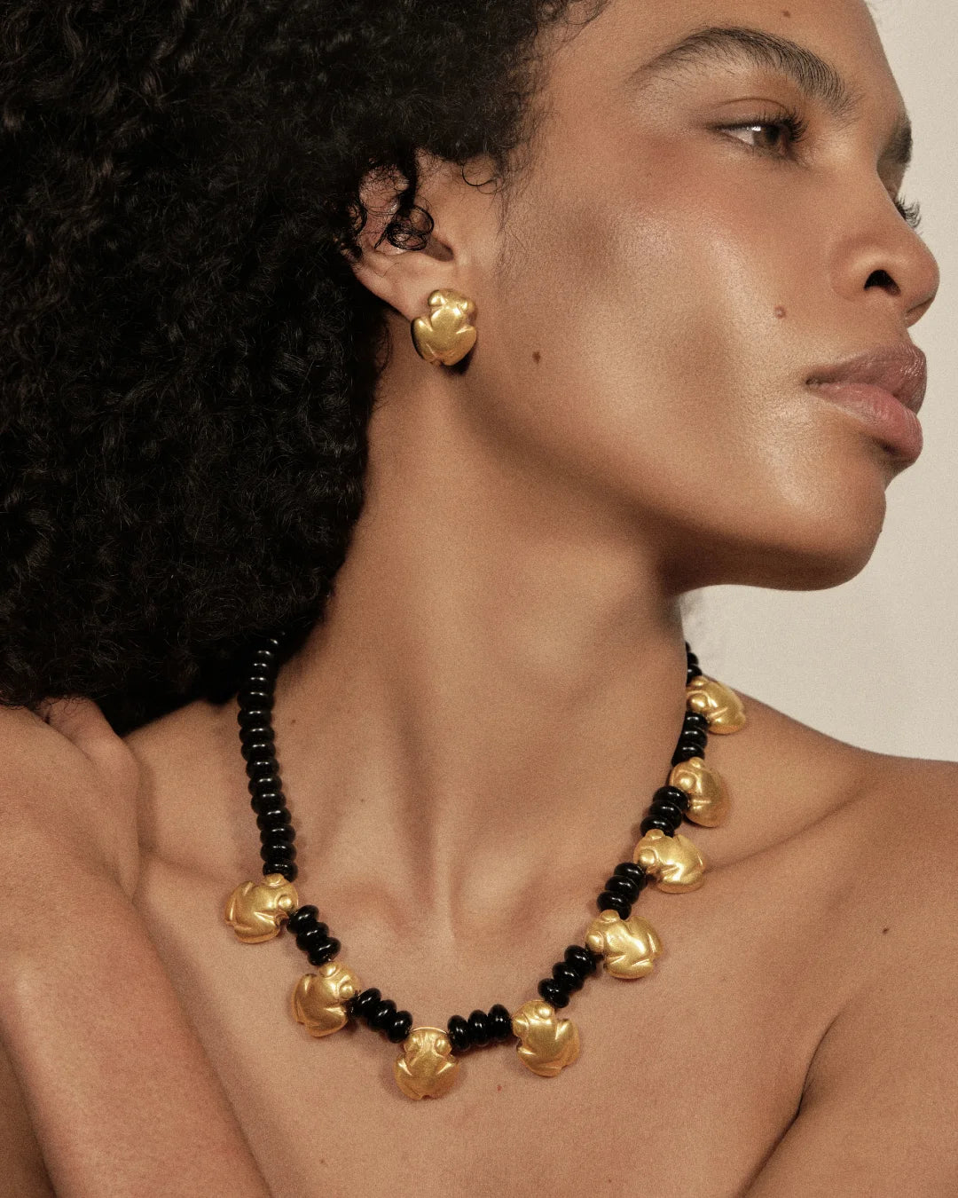 A close-up of a model wearing a statement necklace and matching stud earring. The jewelry features textured, organic-shaped gold beads alternating with small glossy black beads, set against a warm neutral background.