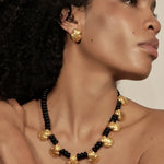A close-up of a model wearing a statement necklace and matching stud earring. The jewelry features textured, organic-shaped gold beads alternating with small glossy black beads, set against a warm neutral background.