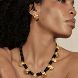 A close-up of a model wearing a statement necklace and matching stud earring. The jewelry features textured, organic-shaped gold beads alternating with small glossy black beads, set against a warm neutral background.